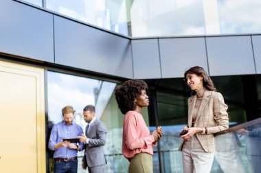 İki kadın çok ırklı meslektaşı ofis dışında arkadaşça sohbet ederken, diğerleri arka planda çalışıyor.