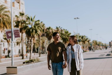 Genç çift Barselona 'da avuçlar tarafından çerçevelenmiş güneşli bir yolda yürürken el ele tutuşup gülümsüyor.