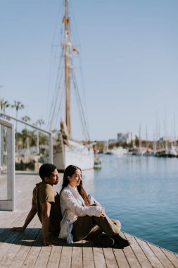 Çift, Barcelona 'da güneşli bir günde limana bakan bir rıhtımda oturup huzurlu bir anın tadını çıkarıyor..