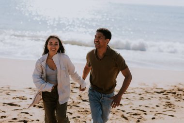 Gülümseyen bir çift kumlu bir sahilde kaygısızca koşmaktan hoşlanır, arka planda yavaşça dalgalar kırılır.