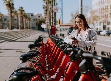 Genç bir kadın güneşli Barcelona 'da bir sıra kırmızı bisikletin yanında duruyor, telefonunu kontrol ediyor, muhtemelen bisiklet paylaşım uygulaması kullanıyor.