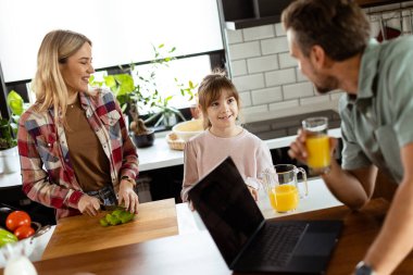 Neşeli bir aile mutfaklarında taze meyve suyu ve masada duran bir dizüstü bilgisayarla sıcak bir sohbetten zevk alır..