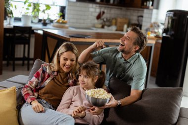 Üç kişilik bir aile rahat bir şekilde kanepeye kurulmuş, heyecanlı bir film gecesinde bir kase patlamış mısırı paylaşırken yüzlerindeki heyecanı ve dikkati yansıtıyor.