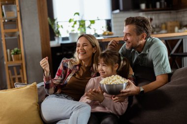 Üç kişilik bir aile rahat bir şekilde kanepeye kurulmuş, heyecanlı bir film gecesinde bir kase patlamış mısırı paylaşırken yüzlerindeki heyecanı ve dikkati yansıtıyor.