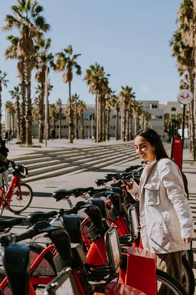 若い女性は晴れたバルセロナで赤い自転車の列に立って 携帯電話をチェックし バイクシェアリングアプリを使用する可能性が高い ストックフォト