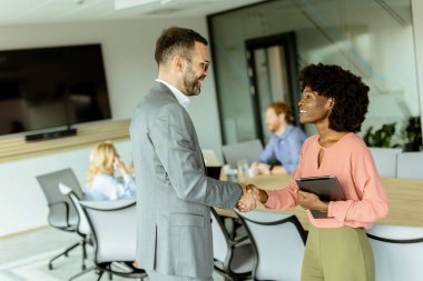 İki profesyonel sıcak bir şekilde el sıkışıyor, anlaşarak gülümsüyor ve takım çalışması arka planda devam ediyor.
