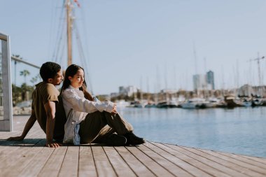 Çift, Barcelona 'da güneşli bir günde limana bakan bir rıhtımda oturup huzurlu bir anın tadını çıkarıyor..