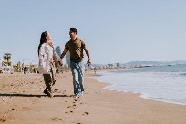 Gülümseyen bir çift kumlu bir sahilde kaygısızca koşmaktan hoşlanır, arka planda yavaşça dalgalar kırılır.