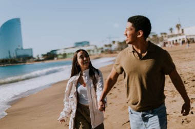 Gülümseyen bir çift kumlu bir sahilde kaygısızca koşmaktan hoşlanır, arka planda yavaşça dalgalar kırılır.