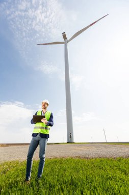 Kask takan bir güvenlik mühendisi, berrak bir gökyüzünün altında yemyeşil ve görkemli bir rüzgar türbininin çalışmalarını inceliyor..