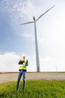 Kask takan bir güvenlik mühendisi, berrak bir gökyüzünün altında yemyeşil ve görkemli bir rüzgar türbininin çalışmalarını inceliyor..