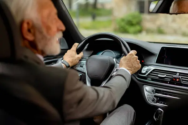 Cheerful elderly man in a suit driving leisurely, basking in the warm glow of a bright day