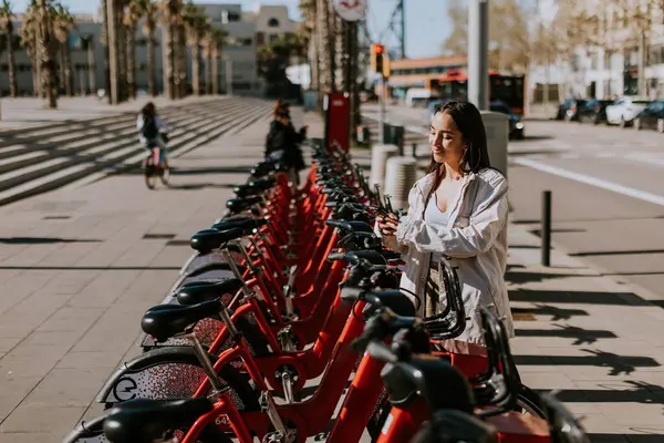 Genç bir kadın güneşli Barcelona 'da bir sıra kırmızı bisikletin yanında duruyor, telefonunu kontrol ediyor, muhtemelen bisiklet paylaşım uygulaması kullanıyor.