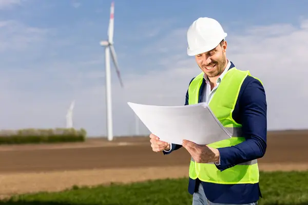 A cheerful man in safety gear scrutinizes blueprints with windmills towering in the background.