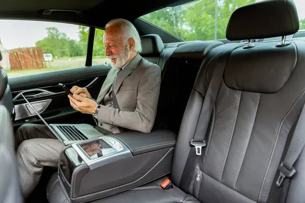 スーツの優れた年上の紳士は 車の乗車中に彼の携帯電話上のメッセージをレビュー 近くの彼のラップトップ ロイヤリティフリーのストック写真