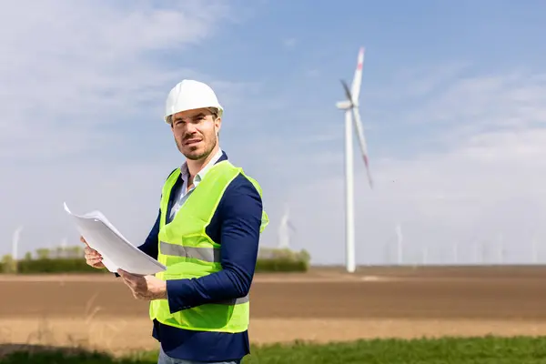 Cheerful Man Safety Gear Scrutinizes Blueprints Windmills Towering Background Royalty Free Stock Images