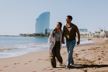 Gülümseyen bir çift kumlu bir sahilde kaygısızca koşmaktan hoşlanır, arka planda yavaşça dalgalar kırılır.