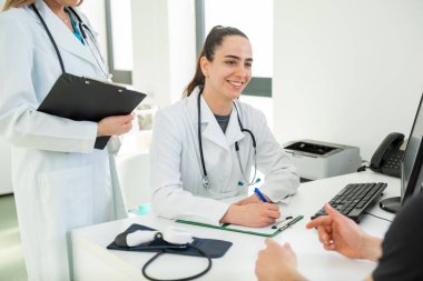 Steteskoplu beyaz önlüklü kadın doktor, bir ofis ortamında notlar alan bir hastaya tavsiye veriyor.