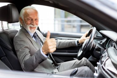 Parlak gülüşlü sakallı yaşlı bir adam, güvenli bir şekilde şehirde arabasını sürerken başparmağını kaldırıyor.