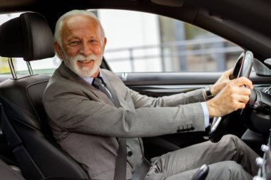 Takım elbise giymiş yaşlı bir beyefendi, güneşli bir günde hareketli bir şehir sokağında modern bir araba sürüyor.