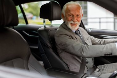 Takım elbise giymiş yaşlı bir beyefendi, güneşli bir günde hareketli bir şehir sokağında modern bir araba sürüyor.