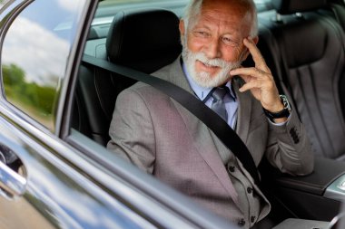 Takım elbiseli zarif bir kıdemli işadamı arabasının arka koltuğundan laptopuna bakarken cep telefonuyla konuşuyor.