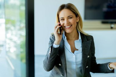 Gri ceketli, kendine güvenen bir iş kadını, parlak bir ofis penceresinin yanında beklerken cep telefonuyla neşeli bir şekilde sohbet ediyor.