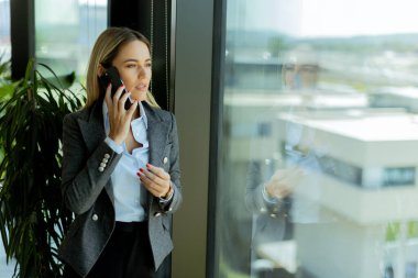 Gri ceketli, kendine güvenen bir iş kadını, parlak bir ofis penceresinin yanında beklerken cep telefonuyla neşeli bir şekilde sohbet ediyor.