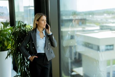 Gri ceketli, kendine güvenen bir iş kadını, parlak bir ofis penceresinin yanında beklerken cep telefonuyla neşeli bir şekilde sohbet ediyor.