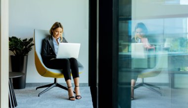 Çağdaş bir ofis salonunda, genç bir profesyonel dizüstü bilgisayarına odaklanıyor, canlı yeşil bir bitkinin yanında, parlak, modern bir sandalyede oturuyor, gün ışığında banyo yapıyor..
