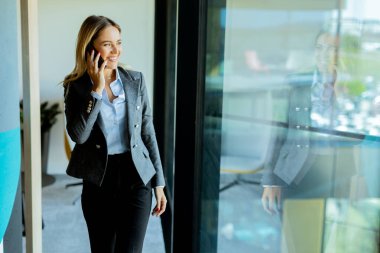 Gri ceketli, kendine güveni tam bir kadın telefonuyla konuşuyor. Çağdaş bir ofis alanında büyük bir pencerenin önünde duruyor ve dışarıdaki güneşli havanın tadını çıkarıyor.