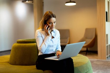 Odaklanmış profesyonel kadın çoklu görevleri verimli bir şekilde telefonda konuşuyor ve bilgisayarında parlak, çağdaş bir ofis alanında daktilo yazıyor.