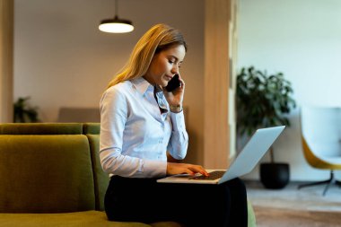 Odaklanmış profesyonel kadın çoklu görevleri verimli bir şekilde telefonda konuşuyor ve bilgisayarında parlak, çağdaş bir ofis alanında daktilo yazıyor.