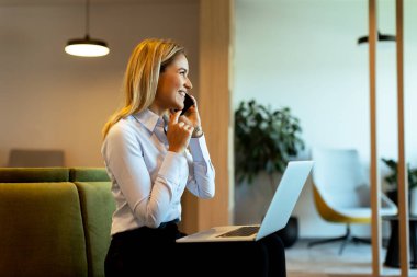 Odaklanmış profesyonel kadın çoklu görevleri verimli bir şekilde telefonda konuşuyor ve bilgisayarında parlak, çağdaş bir ofis alanında daktilo yazıyor.