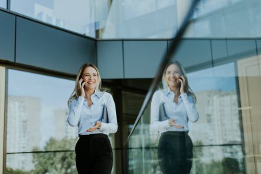 Genç bir iş kadını, zarif, modern bir ofis binasının içinde telefonuyla güvenle gülümsüyor. Camda şehir manzarası yansımaları var..