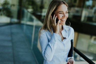 Genç bir iş kadını, zarif, modern bir ofis binasının içinde telefonuyla güvenle gülümsüyor. Camda şehir manzarası yansımaları var..