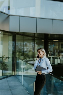 Profesyonel bir iş kadını elinde dizüstü bilgisayarla bir ofis binasının önünde durup gülümsüyor.