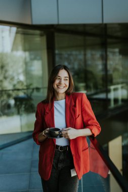 Kırmızı ceket giyen iğrenç kadın elinde kahve fincanı, modern ofis arkaplanı ile dışarıda duruyor, güneşli bir günde pozitiflik ve güven yayıyor.
