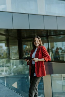 Gülümseyen profesyonel bir kadın, elinde tabletle modern cam ofis binasının yanında duruyor ve akşam boyunca kendinden emin görünüyor..