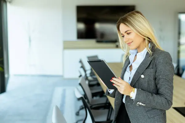 Stock image A cheerful businesswoman in a vibrant red blazer stands confidently in a modern conference room, holding a tablet and ready for a meeting.