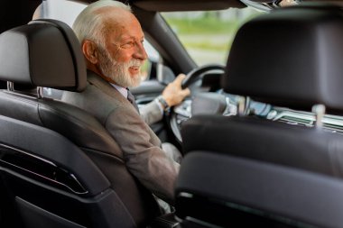 Takım elbise giymiş yaşlı bir beyefendi, güneşli bir günde hareketli bir şehir sokağında modern bir araba sürüyor.