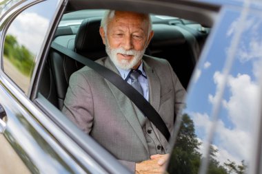 Sakallı, takım elbiseli ve kravatlı yaşlı bir adam emniyet kemeri takmış, aracının içinde mutlu görünüyor.