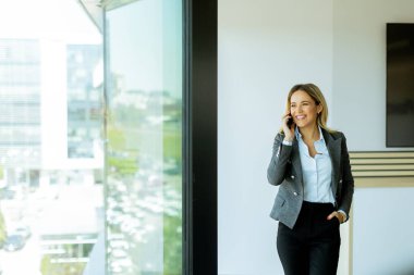 Şık takım elbiseli gülümseyen bir iş kadını cep telefonuyla keyifli bir sohbet ederken dışarıda parlak, güneşli bir manzarası olan büyük bir ofis penceresinin yanında duruyor..