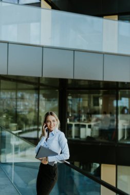 Genç bir iş kadını, zarif, modern bir ofis binasının içinde telefonuyla güvenle gülümsüyor. Camda şehir manzarası yansımaları var..