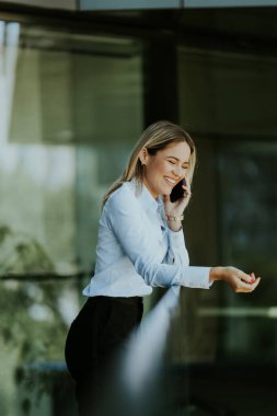 Açık mavi gömlekli, gülümseyen bir kadın, bir korkuluğa dayanır, telefonunu tutar, modern bir ofis binasının önünde, rahatlamanın keyfini çıkarır..