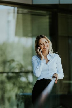 İş kadını, güneşli bir günde, beyaz bluz ve siyah etek giymiş modern bir ofis binasının önünde akıllı telefonuyla konuşurken gülümser.