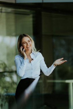 İş kadını, güneşli bir günde, beyaz bluz ve siyah etek giymiş modern bir ofis binasının önünde akıllı telefonuyla konuşurken gülümser.