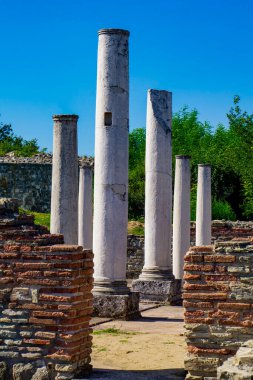 Tall, white columns stand amidst ancient brick ruins in Felix Romuliana, Serbia, with green trees in the background and a clear blue sky overhead clipart