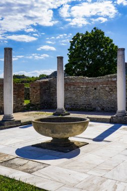 Uzun, beyaz sütunlar Sırbistan 'ın Felix Romuliana kentindeki antik tuğla kalıntılarının arasında duruyor. Arka planda yeşil ağaçlar ve açık mavi bir gökyüzü var.
