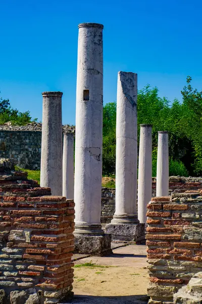 stock image Tall, white columns stand amidst ancient brick ruins in Felix Romuliana, Serbia, with green trees in the background and a clear blue sky overhead